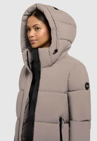 Chaqueta acolchada gris claro con una gran capucha, featuring cremalleras y acentos negros, textura acolchada y logo en la manga.