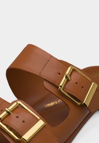 Καφέ δερμάτινο σανδάλι με δύο ρυθμιζόμενες πόρπες από χρυσό μέταλλο και μαξιλαρωτό πέλμα.