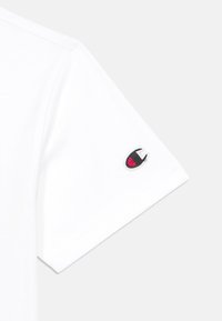 Valge puuvillane T-särk, millel on lühike varrukas ja väike tikandiga logo õmblusjoonel punases, mustas ja valges.