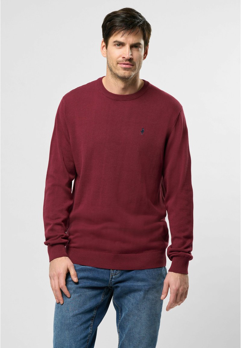 Mann in einem burgunderfarbenen Rundhals-Pullover und blauen Jeans, der vor einem schlichten hellgrauen Hintergrund steht.