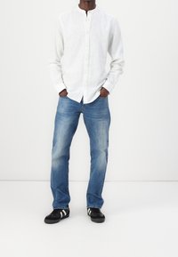 Vit linneskjorta med knappar, blå denimjeans och svarta sneakers med vita ränder. Rakt snitt med ljus tvätt och minimala detaljer.