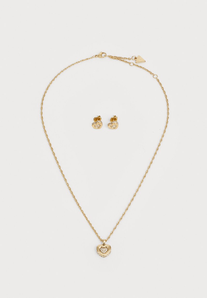 Set di gioielli color oro composto da una collana con catena attorcigliata e un pendente a forma di cuore, abbinata a orecchini a bottone con un piccolo design circolare.