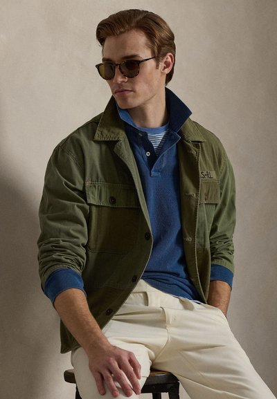 Green cotton jacket over a blue pullover with a striped collar, worn with cream pants and sunglasses, seated against a neutral background.