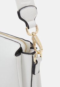 Bílá kožená taška s zlatým zipem a sponou. Černé detaily na okrajích a texturovaný povrch materiálu.