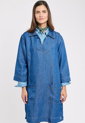 Femme portant une robe en jean bleue jusqu'aux genoux avec de grandes poches, superposée à une chemise bleu clair et un foulard vert autour du cou, debout devant un fond blanc.