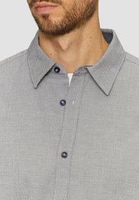 Camicia button-up grigia chiara con motivo a quadretti, tessuto morbido, colletto aperto e bottoni scuri, mostrata nella zona del collo.