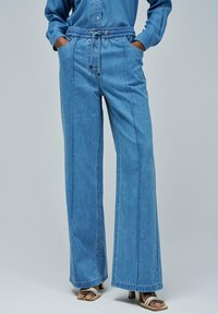 Pantalons en denim à jambe large couleur bleu clair avec une taille élastique, fermeture à cordon et poches frontales. Présente des coutures verticales décoratives.