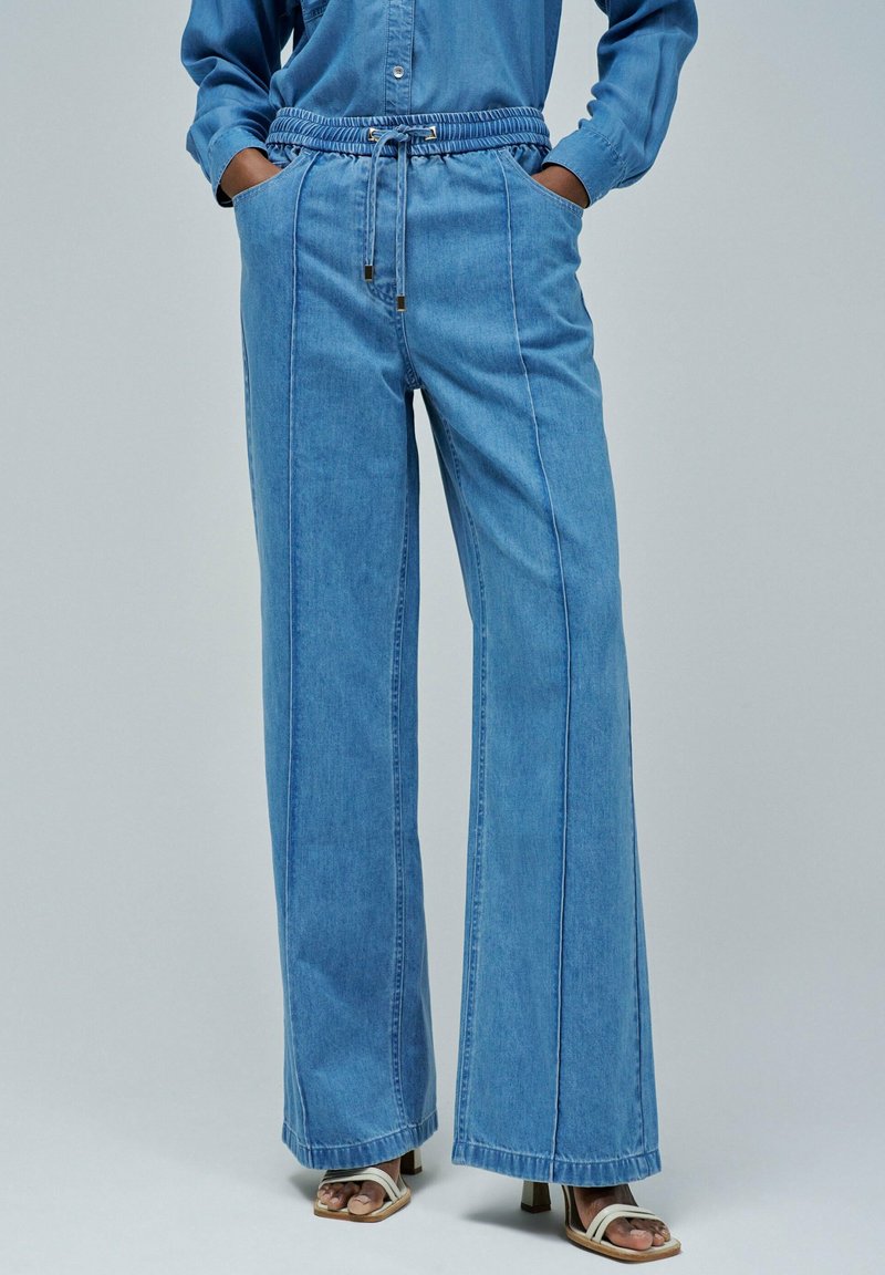 Pantalons en denim à jambe large couleur bleu clair avec une taille élastique, fermeture à cordon et poches frontales. Présente des coutures verticales décoratives.