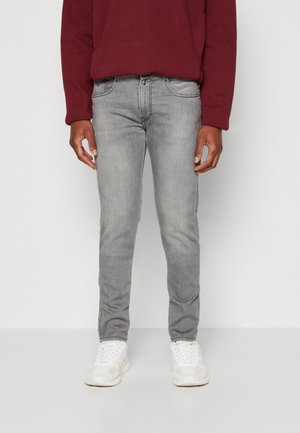 Mand iført slim-fit lysegrå jeans, hvide sneakers og en mørkerød langærmet skjorte, stående mod en ensfarvet lys baggrund.