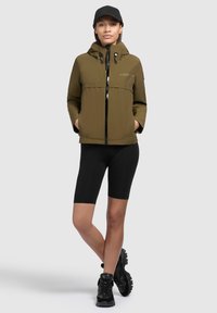 Giacca con cappuccio verde oliva con zip frontale, tasche laterali e dettagli neri, abbinata a pantaloni corti neri e scarpe da ginnastica nere.