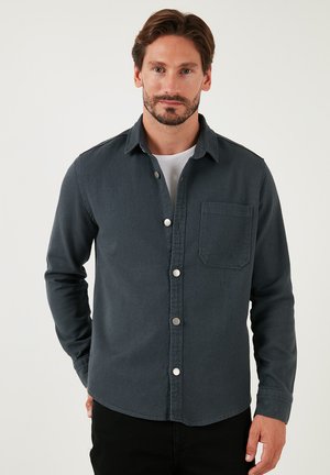 Camisa de botones gris oscuro en un tejido más denso, con un bolsillo en el pecho, mangas largas y botones metálicos. Diseño minimalista con líneas limpias.