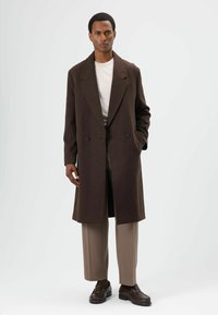 Bruine double-breasted overjas met brede revers, gedragen over een lichtgekleurde ronde hals trui en beige broek, aangevuld met donkere schoenen.