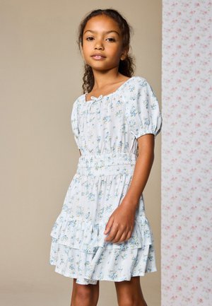 Mädchen in einem weißen Blumen Kleid mit Puffärmeln und einem Stufenrock, das vor einem beigen und geblümten Hintergrund steht.