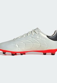 Chaussure de football blanche avec un dessus texturé, design de contour dynamique, accent de talon noir et semelle rouge vif. Comprend six crampons noirs.