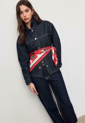 Femme portant une veste en denim foncé et un jean avec une écharpe rouge et blanche à motifs floraux nouée autour de la taille, debout contre un mur clair.