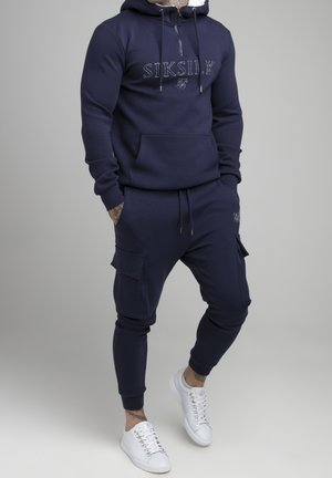 Mann trägt navyblauen Siksilk Hoodie mit Reißverschluss und passenden Cargo-Jogginghosen, kombiniert mit weißen Turnschuhen, steht vor schlichtem Hintergrund.