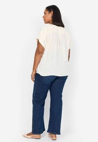 Ljuscremeblus med korta ärmar, flödande design och ryggsöm-detalj, tillsammans med mörkblå utsvängda jeans och beige lägenheter.