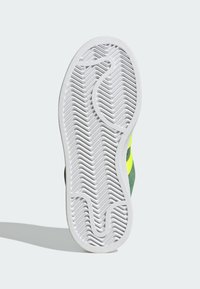 Suela de goma blanca con patrón de relieve en zigzag, parte superior verde con rayas amarillas y detalles texturizados a lo largo de los bordes.