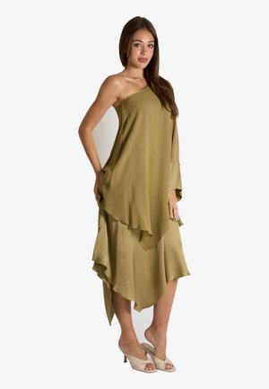 Donna con lunghi capelli castani che indossa un abito asimmetrico monospalla verde oliva e sandali beige con tacco aperto, in piedi su sfondo bianco.