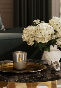 Klar glasskandel med gullakenter, tent veke, plassert på et rundt gullfat. Hvite hortensiaer i en teksturert hvit vase, steinknute skulptur i nærheten.