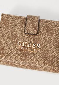 Hnědá texturovaná peněženka s klopou. Obsahuje opakující se vzor loga a zlatý akcent "GUESS EST 1981" na přední straně.