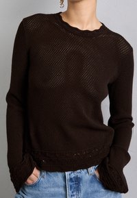 Brauner gestrickter Pullover mit strukturiertem Muster, gewelltem Halsausschnitt und detaillierten Bündchen, kombiniert mit blauen Jeans.
