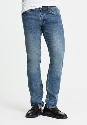 Uomo che indossa jeans di denim blu e mocassini in pelle nera in piedi su uno sfondo bianco semplice.