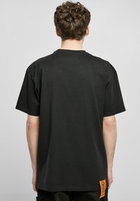 Camiseta negra de manga corta, material de algodón, corte holgado, con una prominente etiqueta de logo naranja cosida en la esquina inferior izquierda.