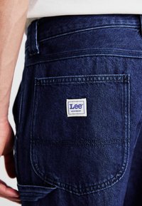Ciemnoniebieskie dżinsy mają wyraźną tylną kieszeń z białą metką z napisem "Lee WORKWEAR". Faktura jest gładka z widocznymi szwami.