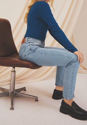 Person trägt blaues Langarmshirt, hellblaue Jeans und schwarze Loafers sitzt seitlich auf einem braunen Drehstuhl vor beige drapiertem Hintergrund.