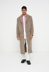 Lång, beige ullblandning kappa med en sjalkrage, två framfickor och en knappstängning. Bärs över en rosa skjorta och vita byxor.