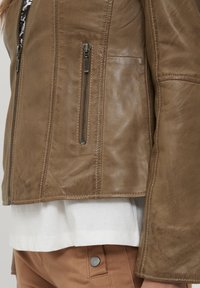 Hnedá kožená bunda so bočnými zipsami, mäkká textúra, navrhnutá s vysokým golierom a mierne rozšíreným rukávom. Preložená cez bielu košeľu.