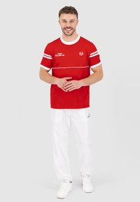 Rode korte mouwen shirt met witte strepen, voorzien van een logo en tekstontwerp, gecombineerd met witte broek. Model staat met handen gevouwen.