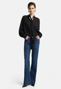 Zwarte blouse met lange mouwen en een strikdetail bij de hals, gecombineerd met donkerblauwe flared jeans en puntige zwarte hakken.