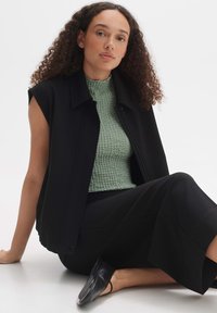 Schwarze ärmellose Oberbekleidung über einem strukturierten grünen Oberteil, kombiniert mit einem schwarzen Rock und schwarzen Ballerinas. Modell sitzt vor einem hellen Hintergrund.