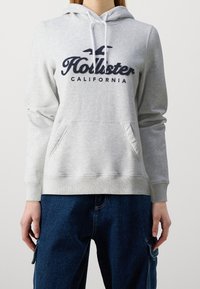Felpa con cappuccio grigio chiaro in tessuto morbido, con tasca a marsupio frontale, cappuccio con coulisse e logo "Hollister California" blu scuro.