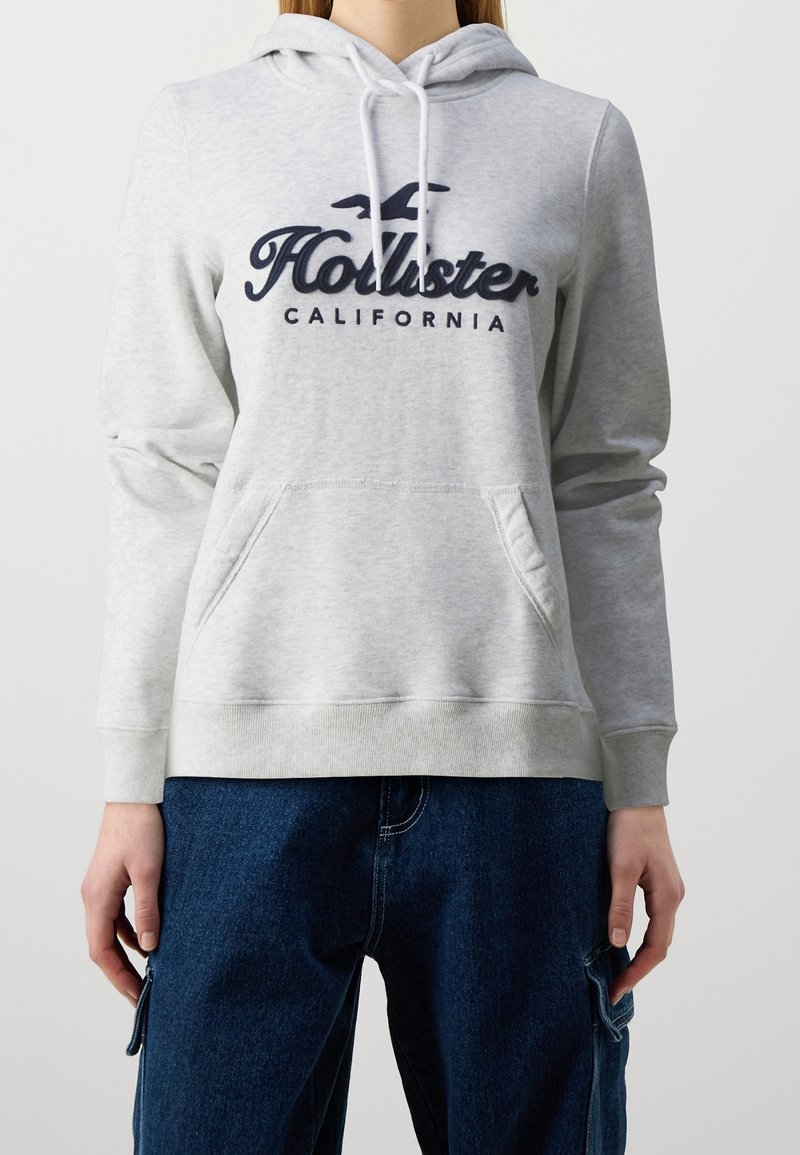 Felpa con cappuccio grigio chiaro in tessuto morbido, con tasca a marsupio frontale, cappuccio con coulisse e logo "Hollister California" blu scuro.