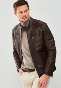 Brown leather motorcycle jacket with a zip closure, shoulder patches, and embroidered logos, worn over a beige sweater and light trousers.