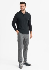 Man står med händerna i fickorna iförd en svart långärmad polo, grå cargobyxor och grå sneakers på en vit bakgrund.