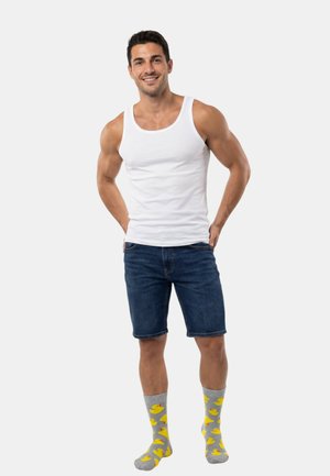 Canotta bianca, pantaloncini di jeans blu, calzini grigi con motivi di paperelle gialle. Modella in piedi con le mani sui fianchi, sorridente contro uno sfondo semplice.