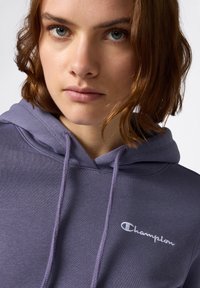 Sudadera morada hecha de una tela suave con un bolsillo tipo canguro en la parte frontal. Presenta una capucha con cordón y el logo de "Champion" bordado en blanco.