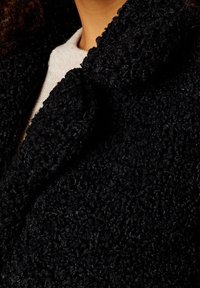 Schwarzer, texturierter Stoff eines Mantels mit einer weichen, fuzzy-artigen Erscheinung, der einen breiten Kragen und ein geschichtetes Design am Ausschnitt aufweist.