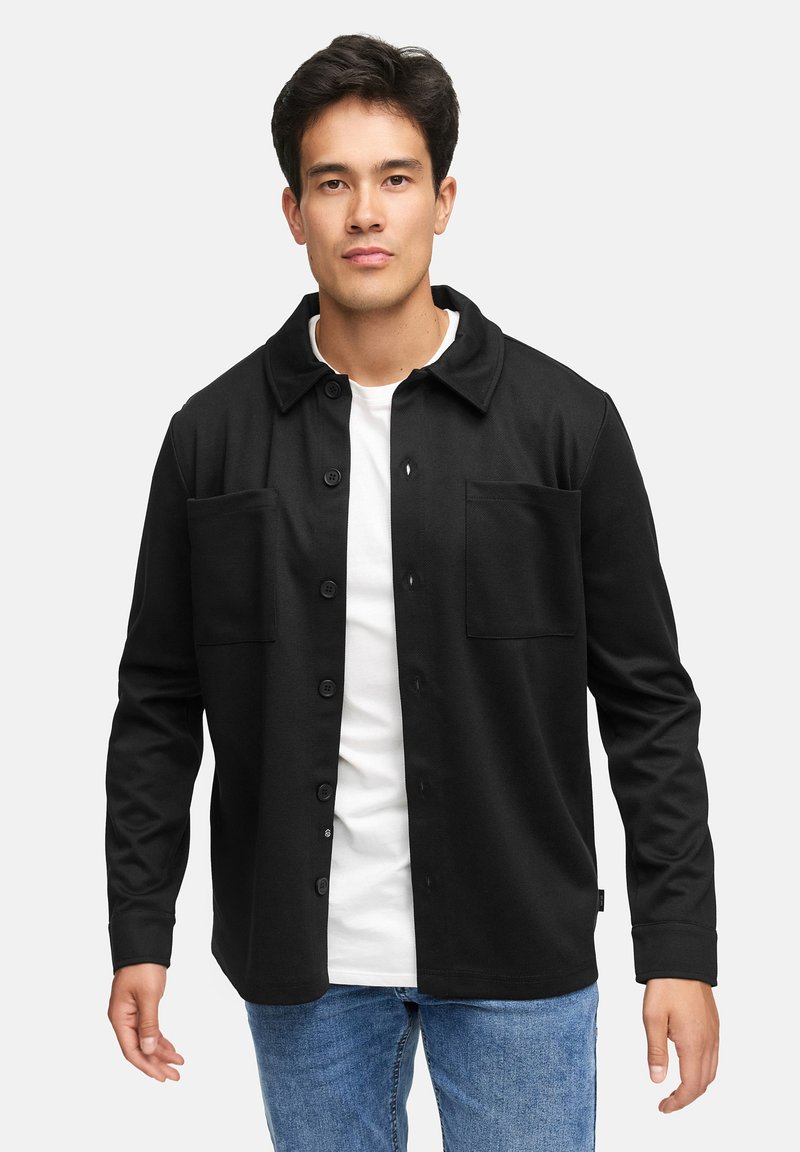 Sort knapp skjorte med krage, to brystlommer og lange ermer, båret over en hvit t-skjorte, kombinert med lyseblå jeans.