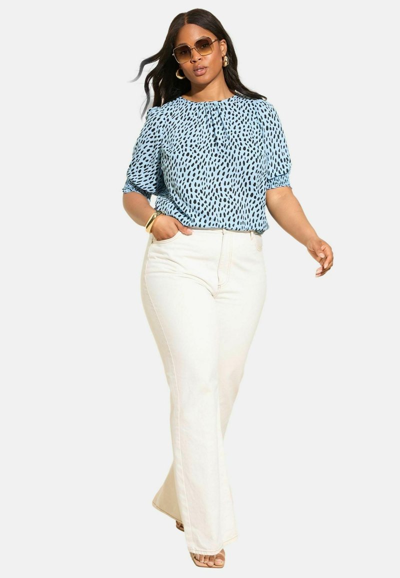 Lysblå bluse med sort prikmønster, elastiske ærmer, parret med højtaljede hvide flarejeans og beige sandaler.