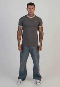 Grå figursydd t-shirt med kontrasterande krämfärgad kant och logotyp, kombinerad med löst sittande blå jeans och vita sneakers. Modellen står mot en neutral bakgrund.