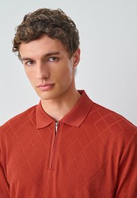 Polo shirt de couleur rouille avec un col zippé, présentant un motif en losanges sur tout le tissu. Matière lisse. Vue de près.
