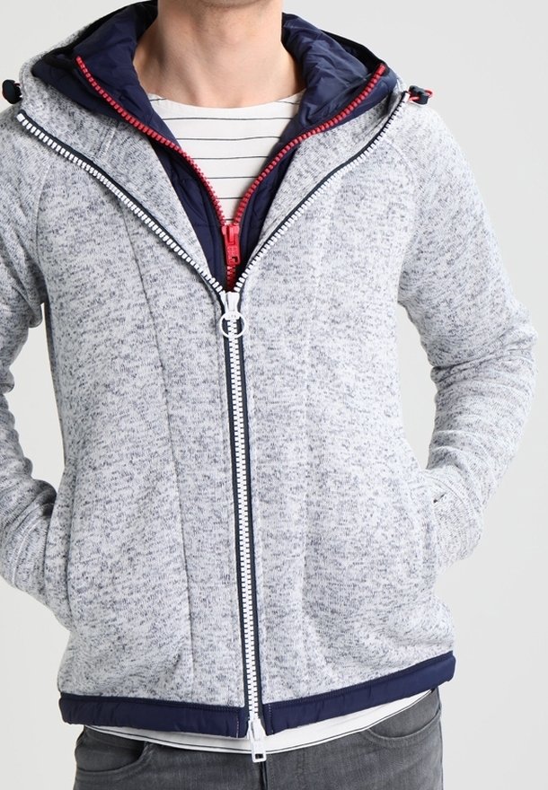 Man som bär flera lager huvor: en grå zip-up hoodie över en marinblå jacka med röd dragkedja, tillsammans med en randig skjorta.