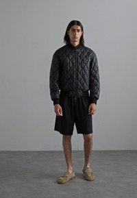 Svart, quiltad bomberjacka med dragkedja, bärs över svarta shorts och tan färgade slip-on skor. Enfärgad bakgrund och medelgrå golvtextur.