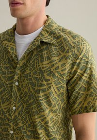 Camicia a maniche corte in tessuto verde e giallo con un motivo astratto, chiusura a bottoni e colletto all'americana. Texture morbida e design casual.