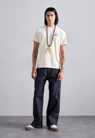 Cream t-shirt with logo, loose dark denim pants with orange stitching, brown beaded necklace with tassel, and white sneakers.